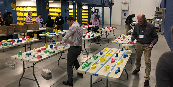Workshop attendees carry colored blocks around multiple tables in a large room.