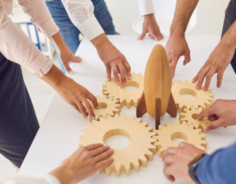 A team's hands reaching toward a set of wooden gears and rocket sitting on a table. 