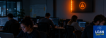 A dark software team room or open office, with a single wall monitor or andon light glowing Orange while the people nearby keep working, heads down. The light is plainly visible and plainly unanswered.